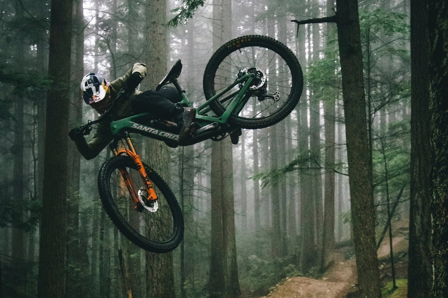 Santa Cruz green and orange Bike flying through the forest with both wheels in the air.